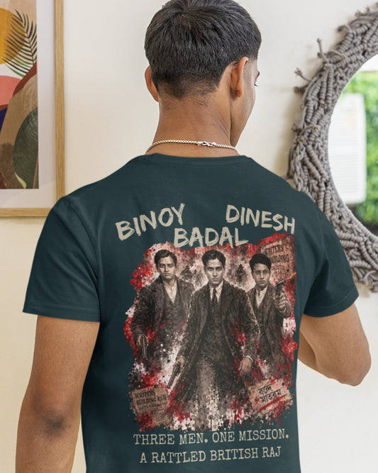 A man viewed from behind wearing a dark teal-green t-shirt with a large graphic print on the back. The print features the text 'BINOY DINESH BADAL' and 'THREE MEN, ONE MISSION, A RATTLED BRITISH RAJ' in white, surrounding a vintage-style illustration of three men in suits with red blood splatter details.