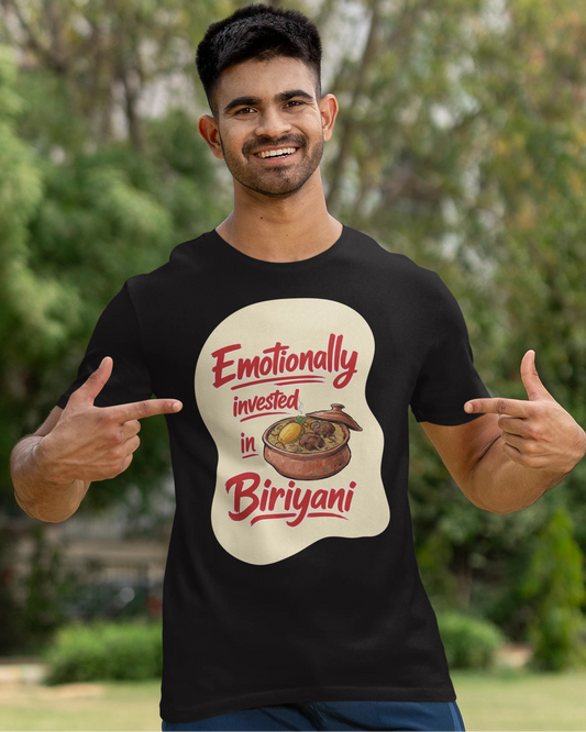 A man wearing a black graphic t-shirt with a 'Emotionally invested in Biriyani' print, posing outdoors with a cricket bat against a blurred green background.