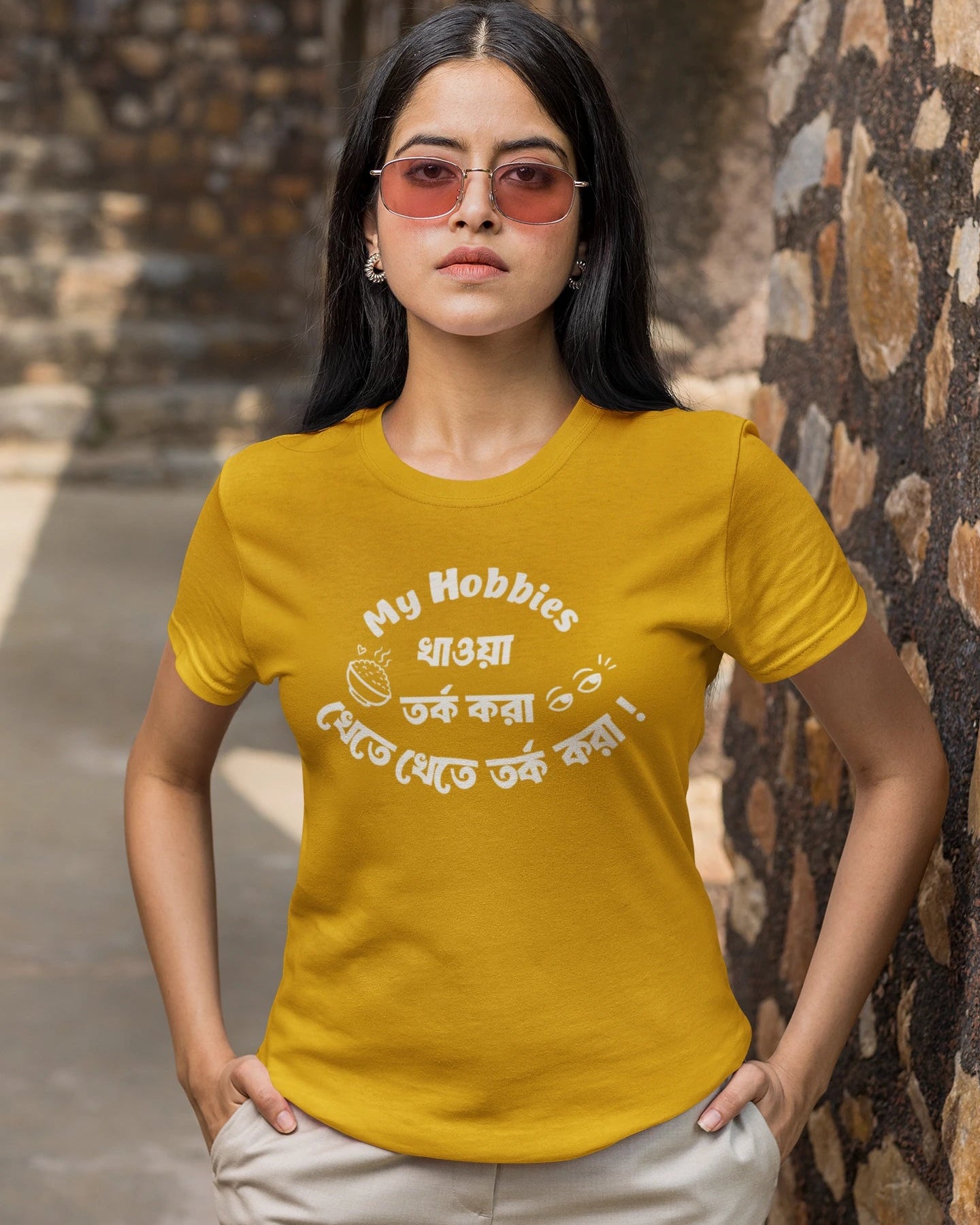 A woman wearing a mustard yellow graphic t-shirt with white text in English and Bengali, paired with beige pants, standing outdoors against a rustic stone wall.