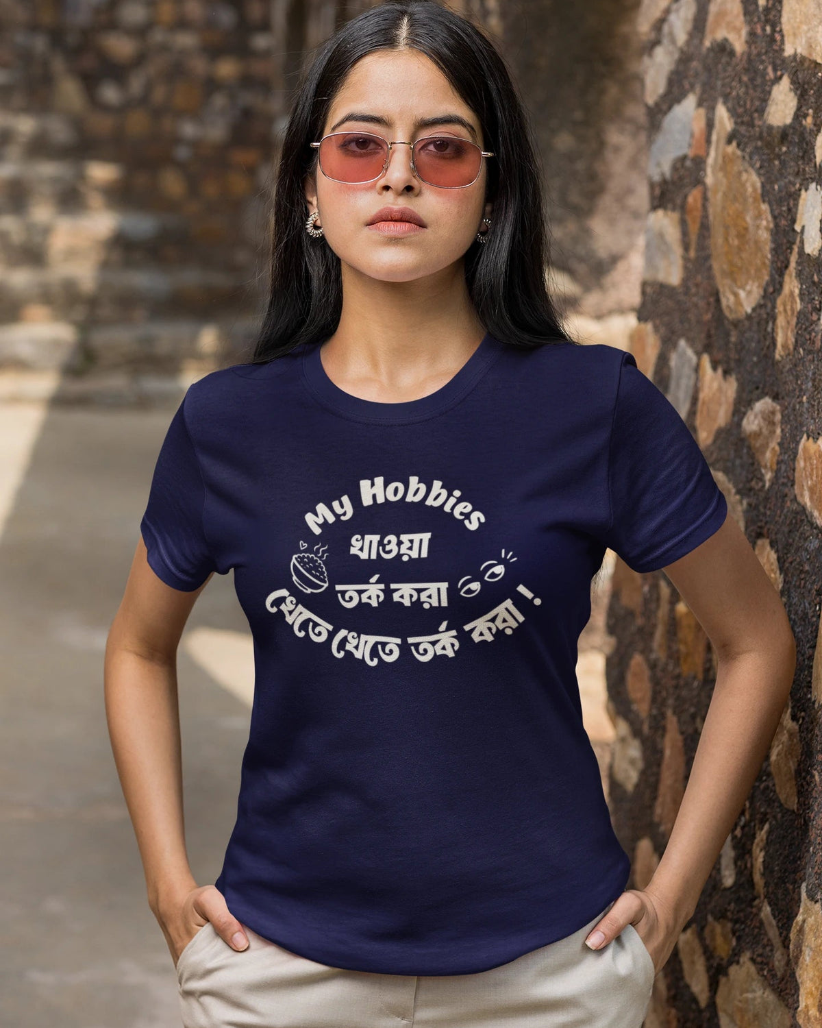 A woman wearing a navy blue t-shirt with white Bengali text and a coffee cup graphic, paired with beige pants, standing against a rustic stone wall.