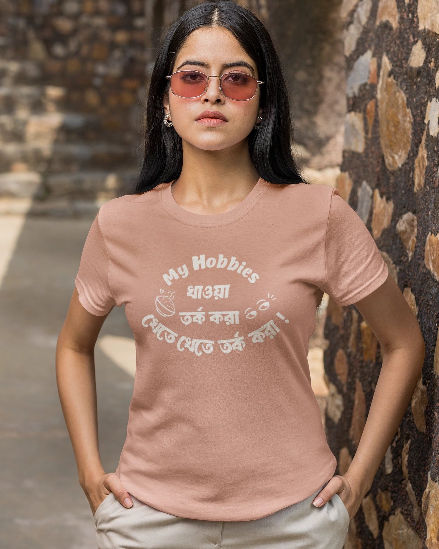 A woman wearing a dusty rose pink t-shirt with a white graphic print that reads 'My Hobbies খাওয়া দেখতে তক করো!' in English and Bengali, paired with beige pants and pink-tinted sunglasses, standing against a rustic stone wall.