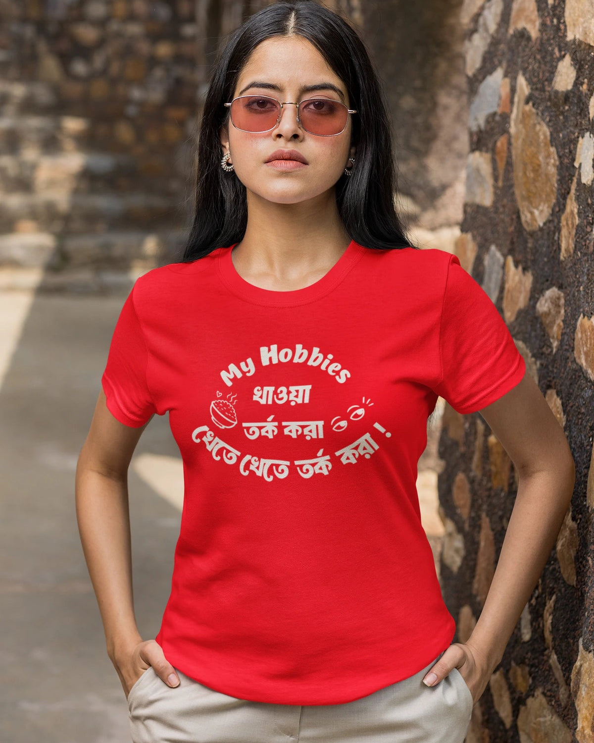 A woman wearing a bright red graphic t-shirt with white Bengali text and a coffee cup illustration, paired with beige pants, standing outdoors against a stone wall.