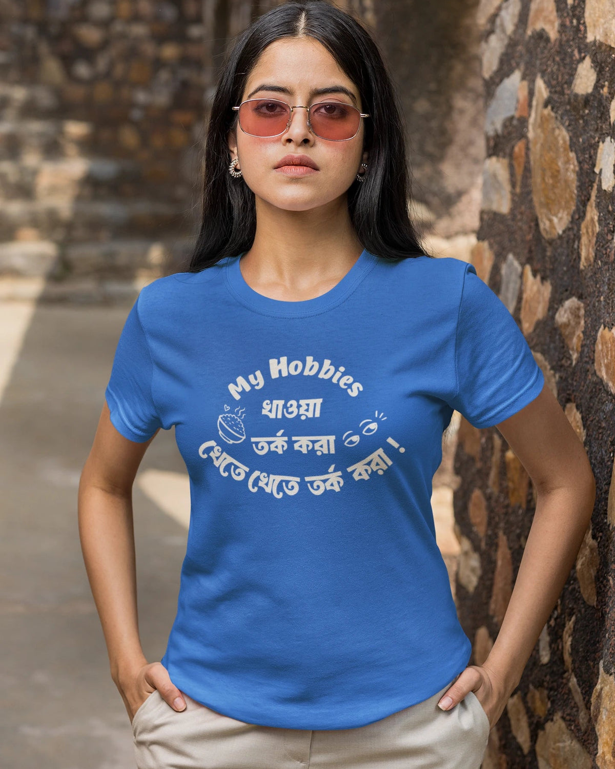 A woman wearing a royal blue t-shirt with a white graphic print that reads 'My Hobbies ধারা তর্ক করা থেকে যত্বে তর্ক করা' and beige pants, standing outdoors against a rustic stone wall.