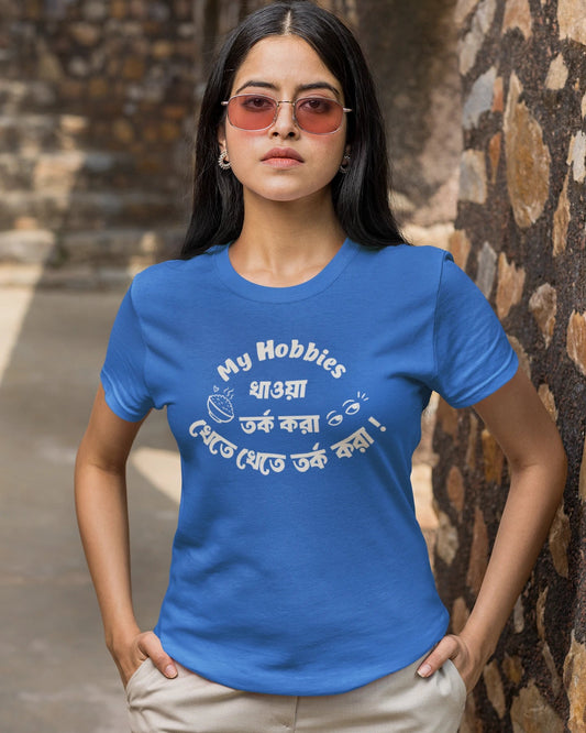 A woman wearing a royal blue t-shirt with a white graphic print that reads 'My Hobbies ধারা তর্ক করা থেকে যত্বে তর্ক করা' and beige pants, standing outdoors against a rustic stone wall.