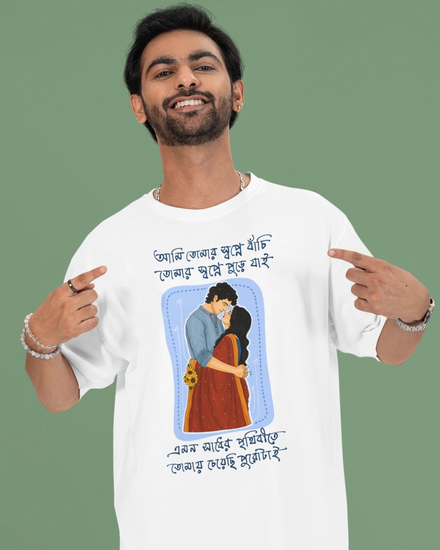 A man wearing a white graphic t-shirt with a Bengali illustration and text, pointing at the design on the front. The background is a solid sage green.