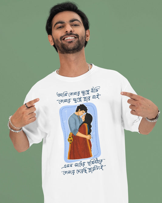 A man wearing a white graphic t-shirt with a Bengali illustration and text, pointing at the design on the front. The background is a solid sage green.