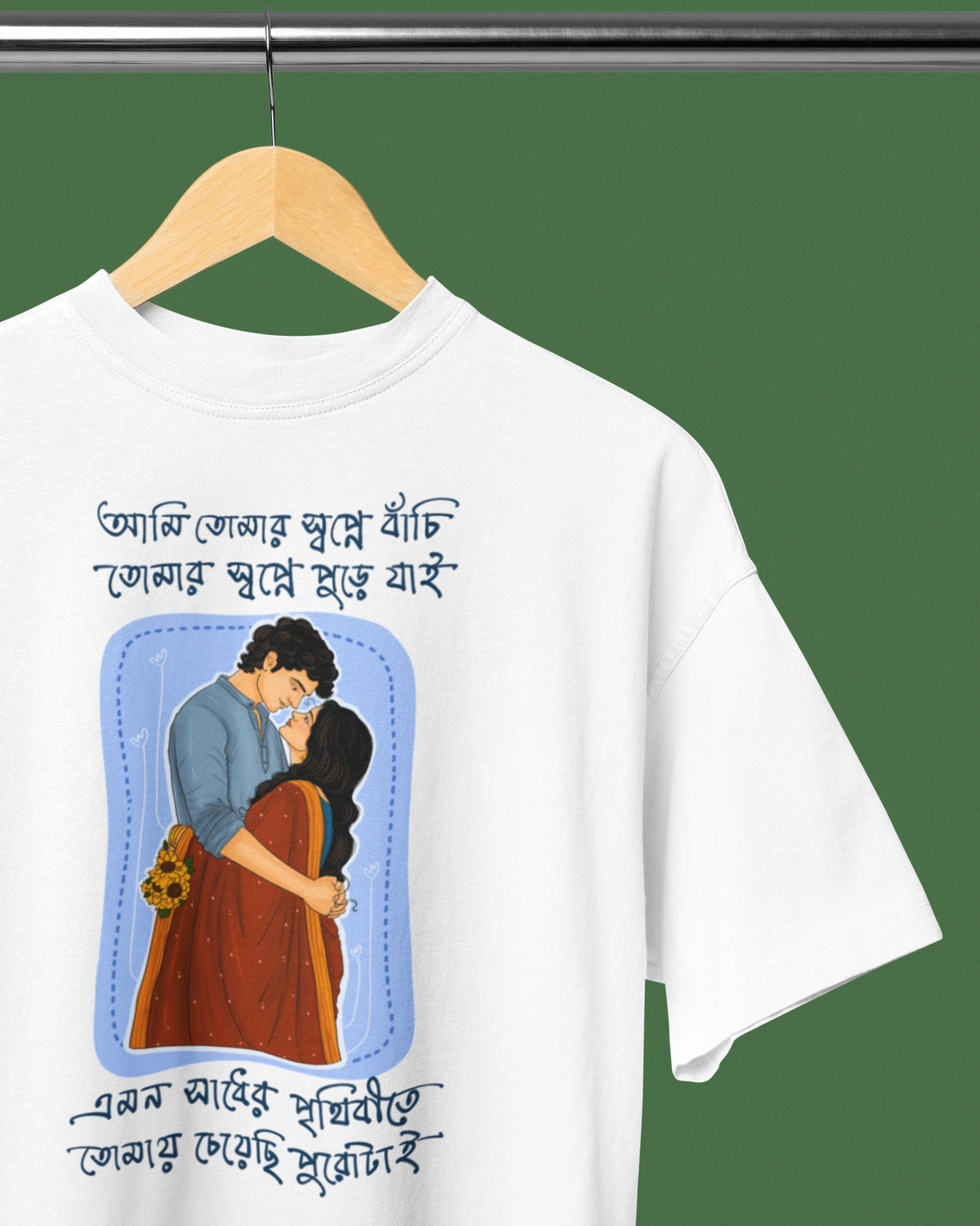 A white short-sleeve t-shirt hanging on a wooden hanger against a solid dark green background. The shirt features a graphic print of a couple embracing, surrounded by blue text in Bengali script.