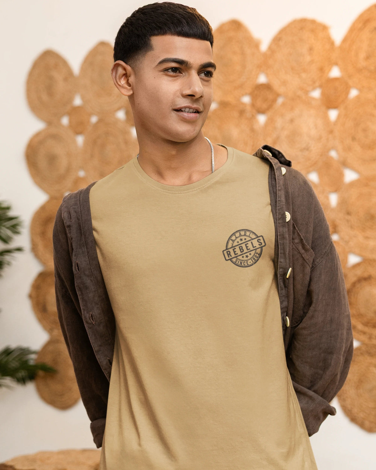 A man wearing a beige crew-neck t-shirt with a circular chest graphic, layered with a dark brown button-up shirt, standing in front of a decorative wooden wall.