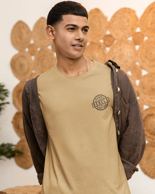A man wearing a beige crew-neck t-shirt with a circular chest graphic, layered with a dark brown button-up shirt, standing in front of a decorative wooden wall.