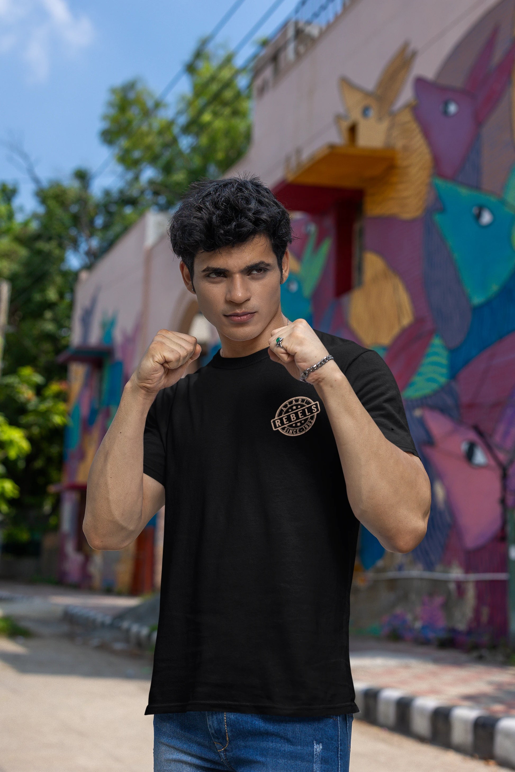 A man wearing a black crew-neck t-shirt with a circular gold graphic on the left chest, posing with his fists raised in front of a colorful graffiti wall.