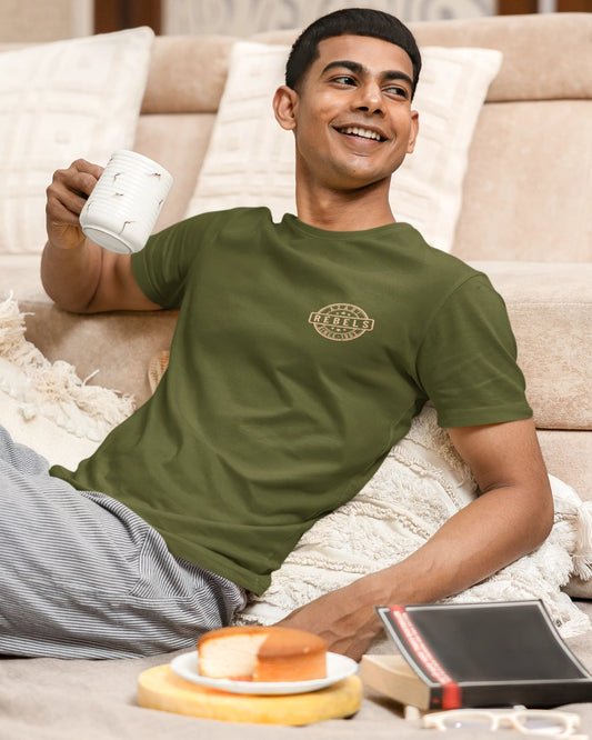 A man wearing an olive green crew-neck t-shirt and gray striped pajama pants relaxes on a beige sofa, holding a white mug. A small circular logo is printed on the left chest of the t-shirt.