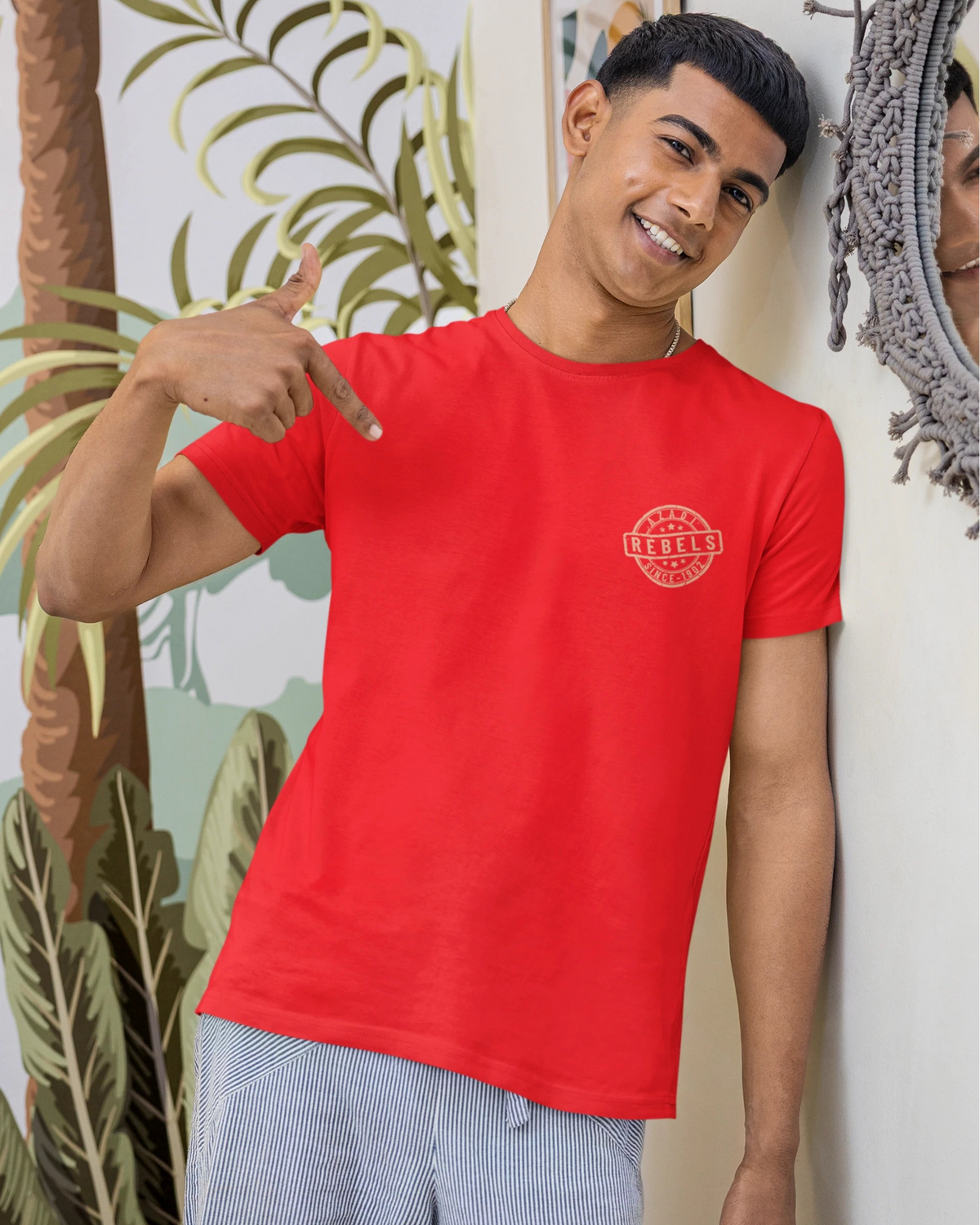 A man wearing a bright red crewneck t-shirt with a circular red graphic on the left chest, paired with light gray striped pajama pants, posing in front of a tropical leaf wallpaper and a decorative mirror.