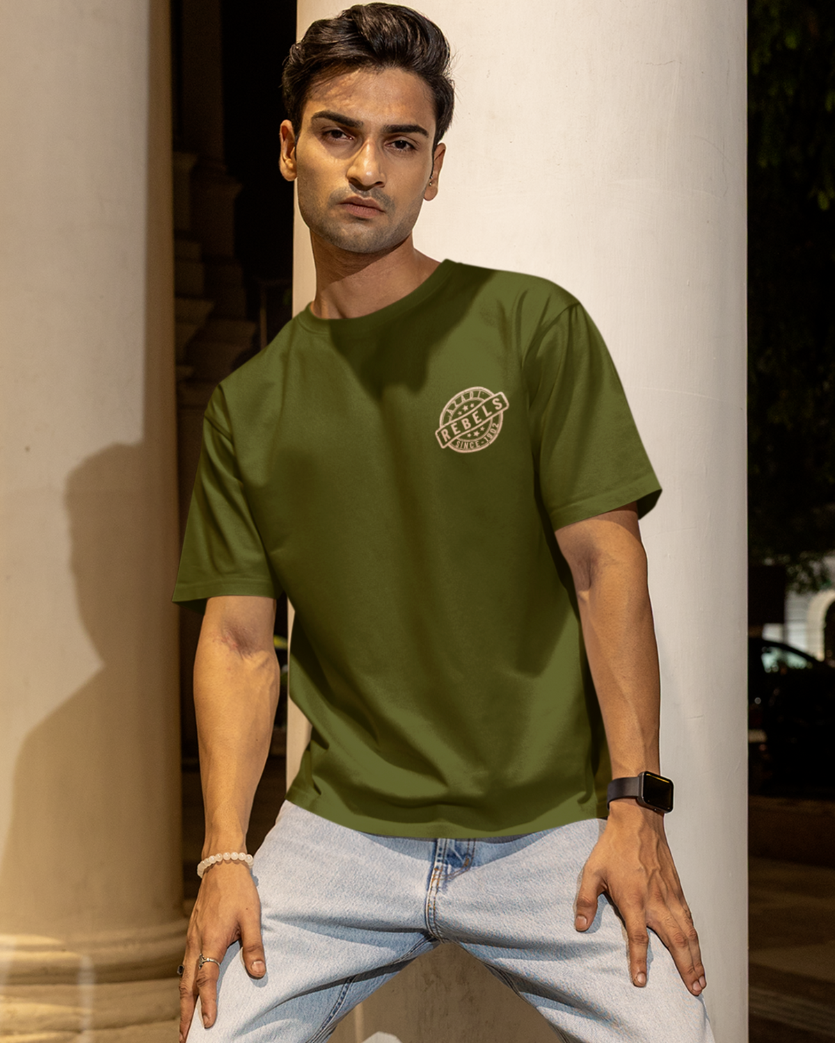 A man wearing an olive green crewneck t-shirt with a small circular graphic on the left chest, paired with light-wash jeans, posing in front of a large white stone column.