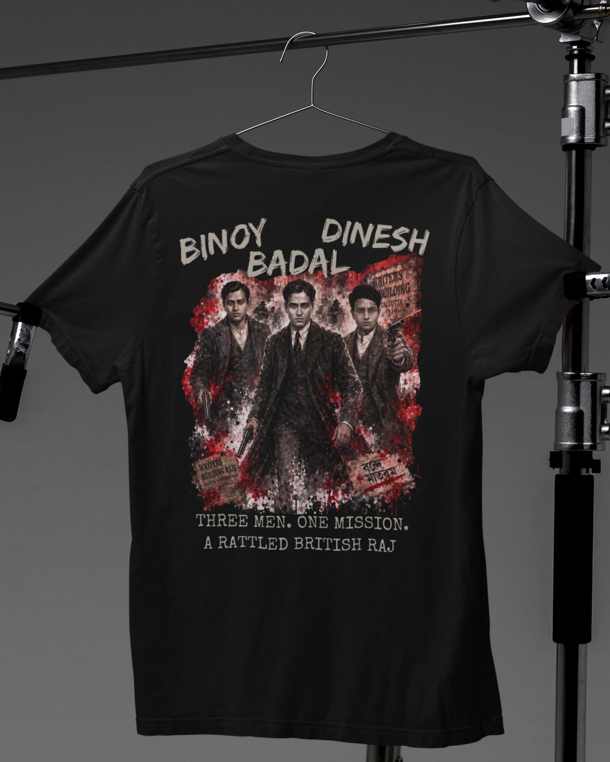 A black short-sleeve T-shirt hanging on a hanger against a dark gray studio background, with a large front graphic print featuring the text 'BINAY DINESH BADAL' and a distressed red-and-white illustrated scene with three men in suits. The shirt is photographed between black studio stands.