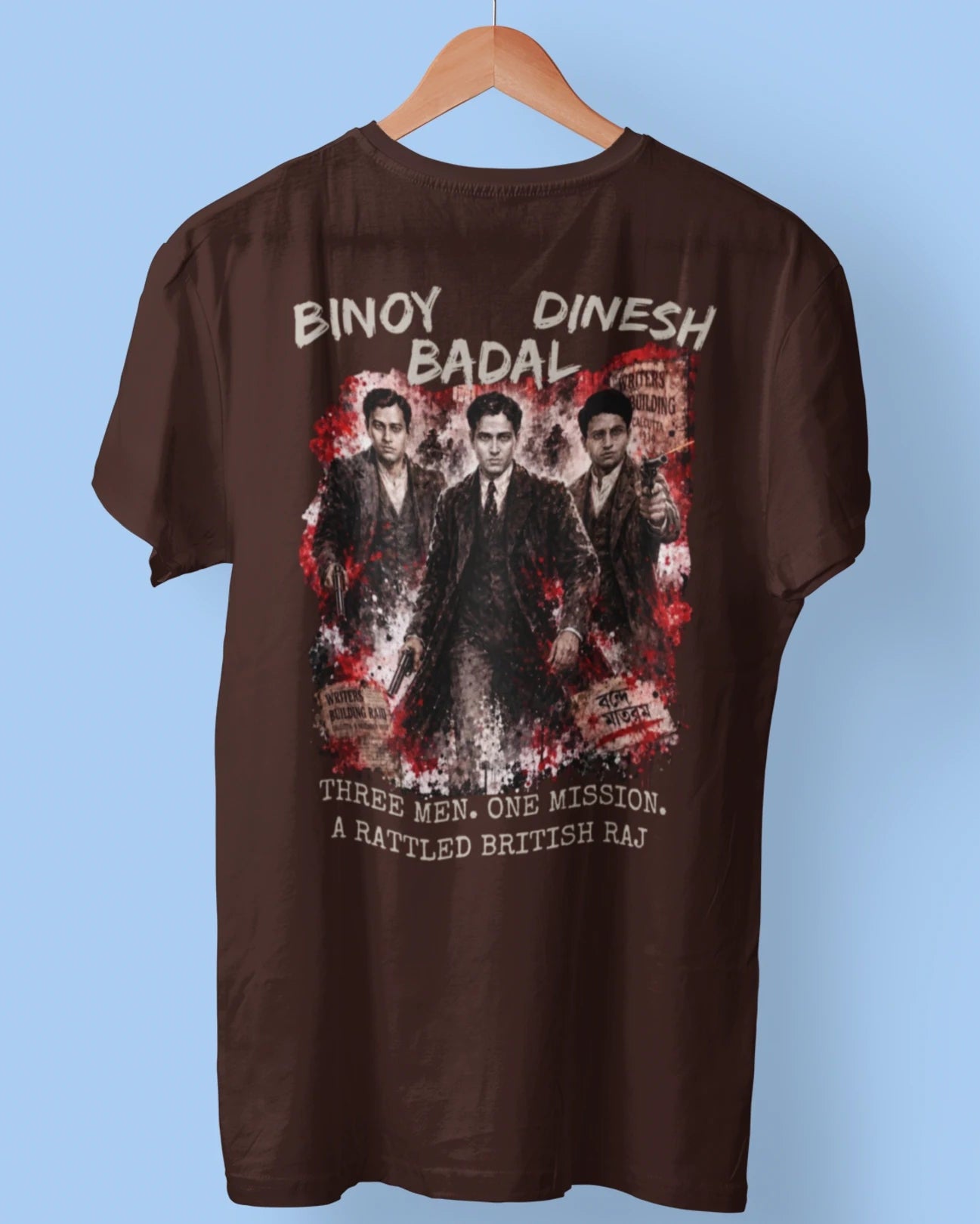 A brown short-sleeve t-shirt hanging on a wooden hanger against a light blue background. The back of the shirt features a large graphic print of three men in suits with red and white distressed splatter details, accompanied by the text 'BINAY DINESH BADAL' and 'THREE MEN, ONE MISSION, A BATTLED BRITISH Raj'.