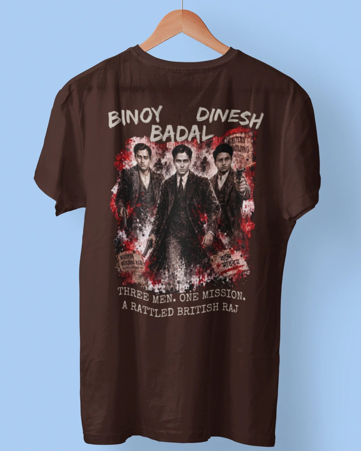A brown short-sleeve t-shirt hanging on a wooden hanger against a light blue background. The back of the shirt features a large graphic print of three men in suits with red and white distressed splatter details, accompanied by the text 'BINAY DINESH BADAL' and 'THREE MEN, ONE MISSION, A BATTLED BRITISH Raj'.