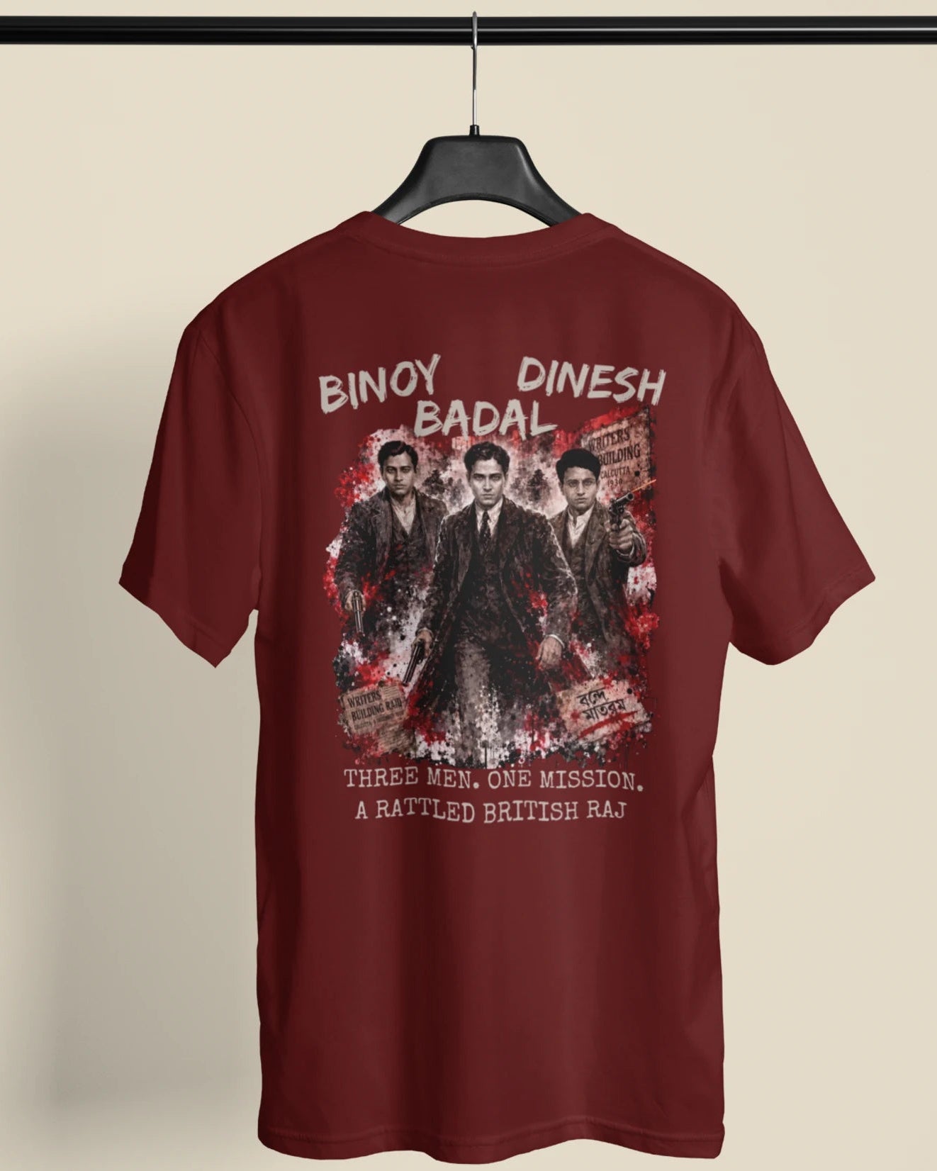 A maroon short-sleeve t-shirt hanging on a black hanger against a light beige background. The back of the shirt features a large graphic print of three men in suits with white text reading 'BINAY DINESH BADAL' and 'THREE MEN, ONE MISSION, A BATTLED BRITISH Raj'.