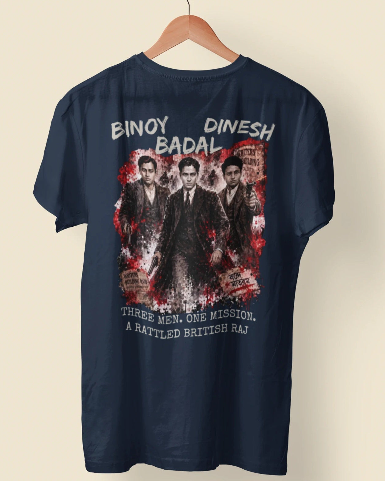 A navy blue short-sleeve t-shirt hanging on a wooden hanger against a light beige background. The back of the shirt features a large graphic print with the text 'BINAY DINESH BADAL' at the top and 'THREE MEN, ONE MISSION, A BATTLED BRITISH RAJ' at the bottom, surrounding a central image of three men in suits.