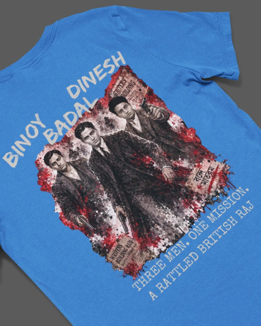 A bright blue short-sleeve t-shirt laid flat on a dark gray background, featuring a large graphic print on the back with the text 'BINAY DINESH' and a distressed black-and-white photo of three men with red splatter accents and additional text below.
