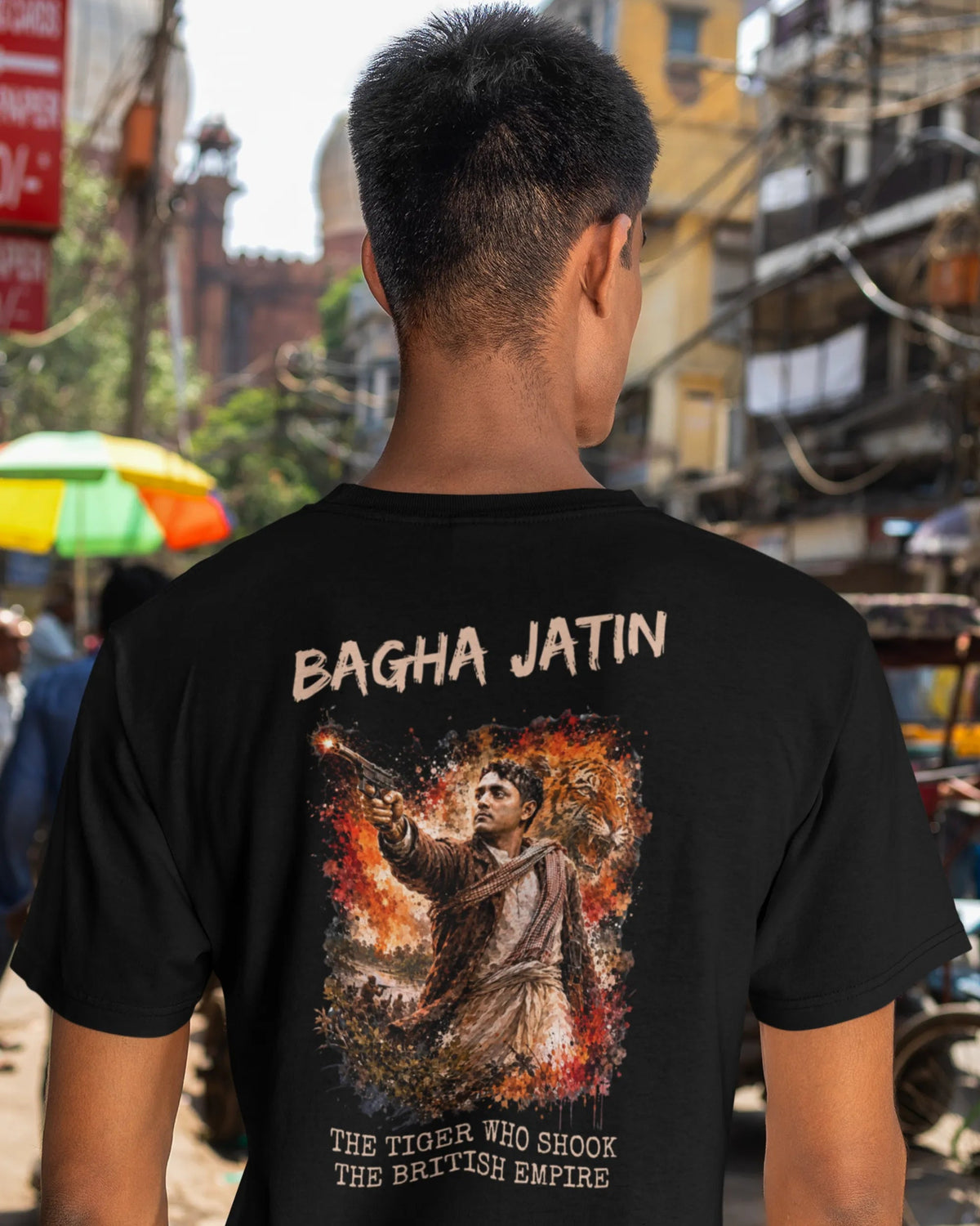 A man viewed from behind wearing a black t-shirt with a large graphic print of Bagha Jatin from the movie Excess Baggage. The street background is blurred.