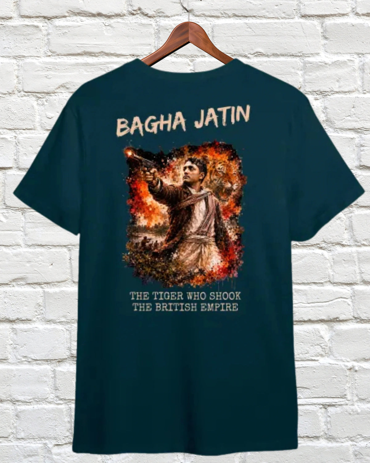 A dark teal t-shirt hanging on a wooden hanger against a white brick wall. The back of the shirt features a large graphic print of a tiger surrounded by flames, with the text 'BAGHA JATIN' above and 'THE TIGER WHO SHOOK THE BRITISH EMPIRE' below.