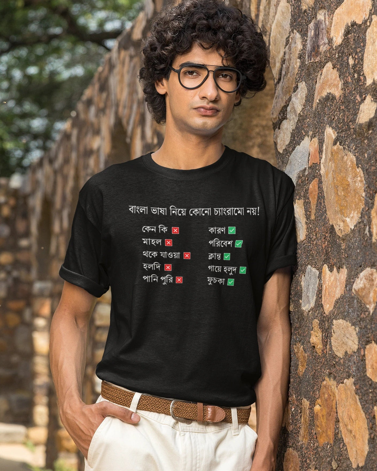 A man wearing a black graphic t-shirt with Bengali text and red and green symbols, paired with white pants and glasses, standing against a rustic stone wall.