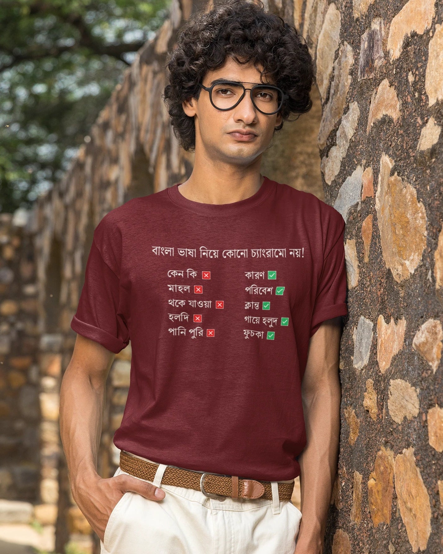 A man with curly black hair and glasses wearing a maroon t-shirt with Bengali text and white pants, standing outdoors against a stone wall.