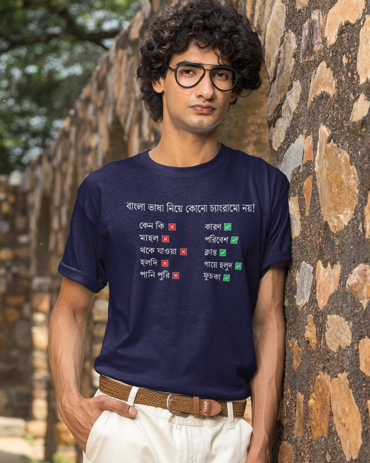A man wearing a navy blue graphic t-shirt with Bengali text and a list of names in red, green, and white, paired with off-white trousers and glasses, standing against a rustic stone wall.