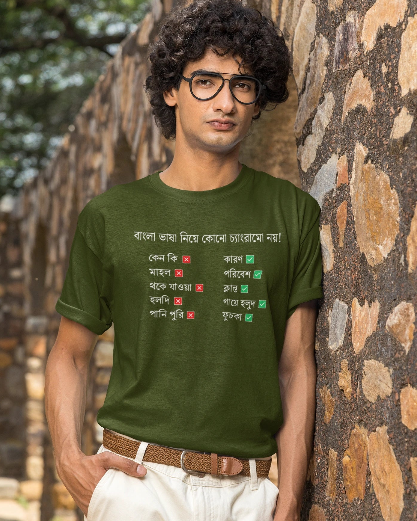 A man wearing a dark green graphic t-shirt with Bengali text, white pants, and glasses, standing outdoors against a rustic stone wall.