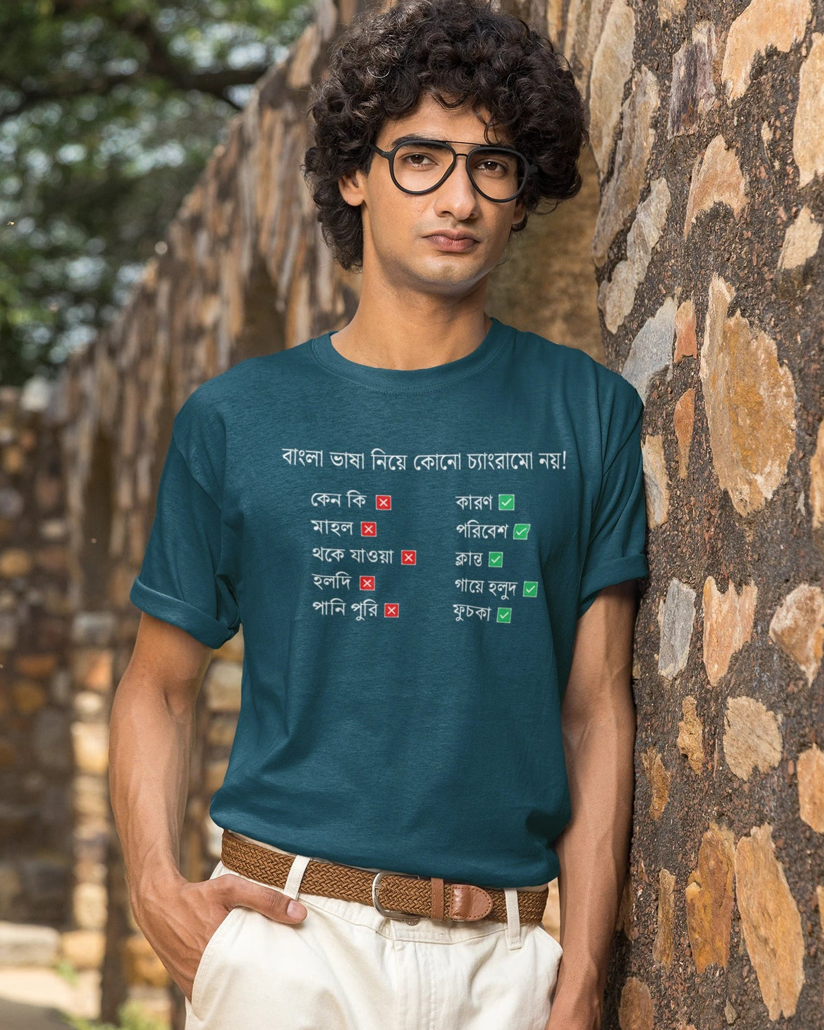 A man wearing a teal blue graphic t-shirt with Bengali text, paired with off-white trousers and glasses, standing outdoors against a rustic stone wall.