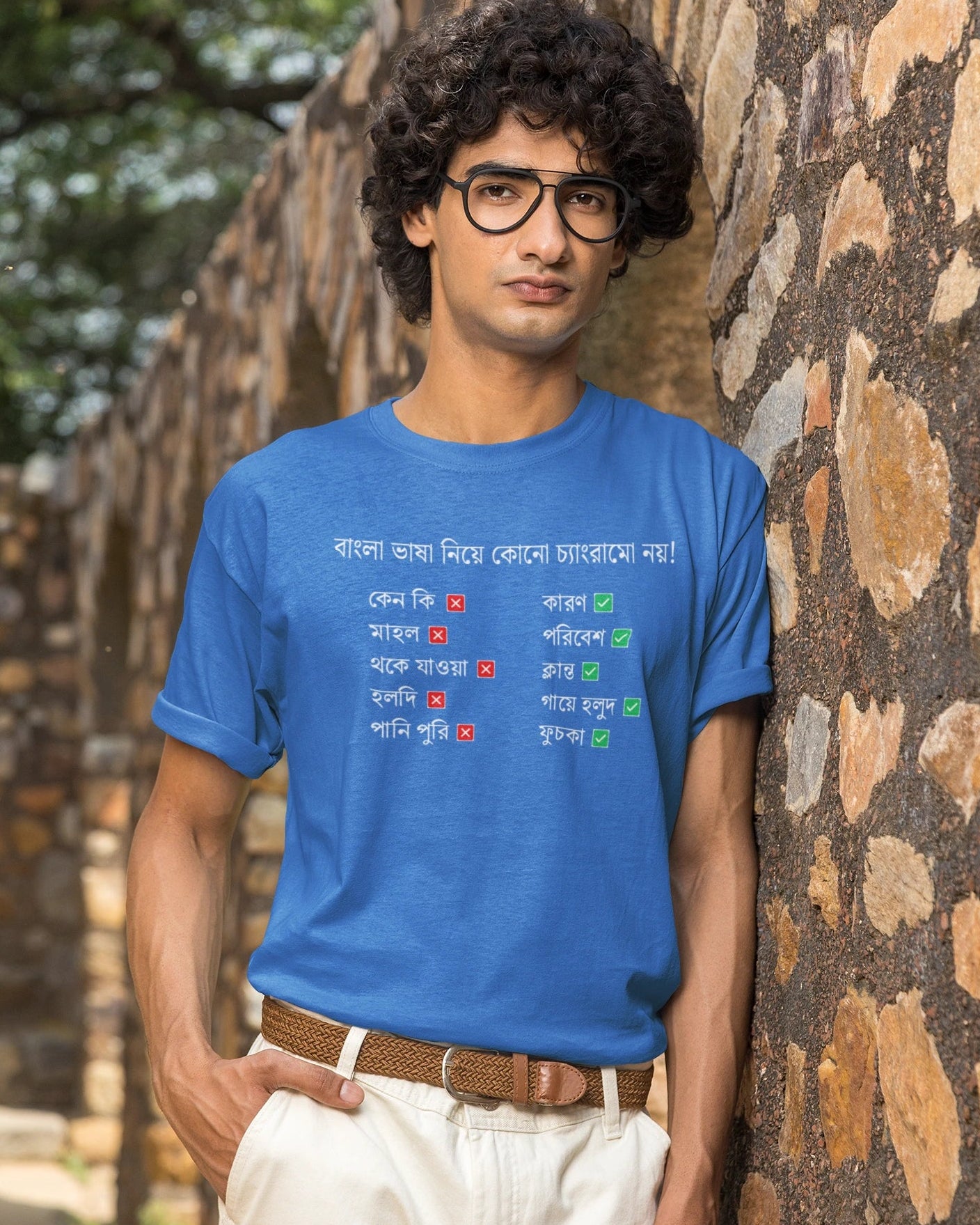 A man wearing a bright blue graphic t-shirt with Bengali text and red/green checkmarks, paired with off-white trousers and glasses, standing outdoors against a rustic stone wall.