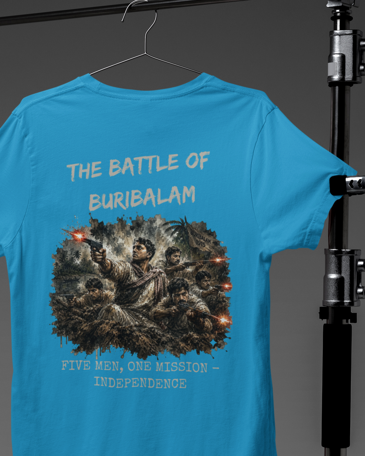 A bright blue t-shirt hanging on a hanger, showing the back side with a detailed graphic print and text reading 'THE BATTLE OF BURIBALAM' and 'FIVE MEN, ONE MISSION - INDEPENDENCE'. The shirt is photographed against a dark gray background with studio lighting equipment visible on the right side.