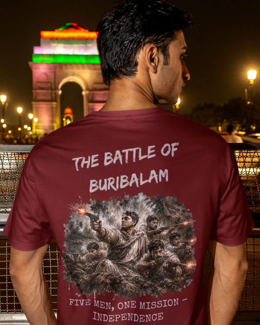 A man viewed from behind wearing a maroon t-shirt with a graphic print of the Battle of Burubalam on the back, standing in front of India Gate at night.