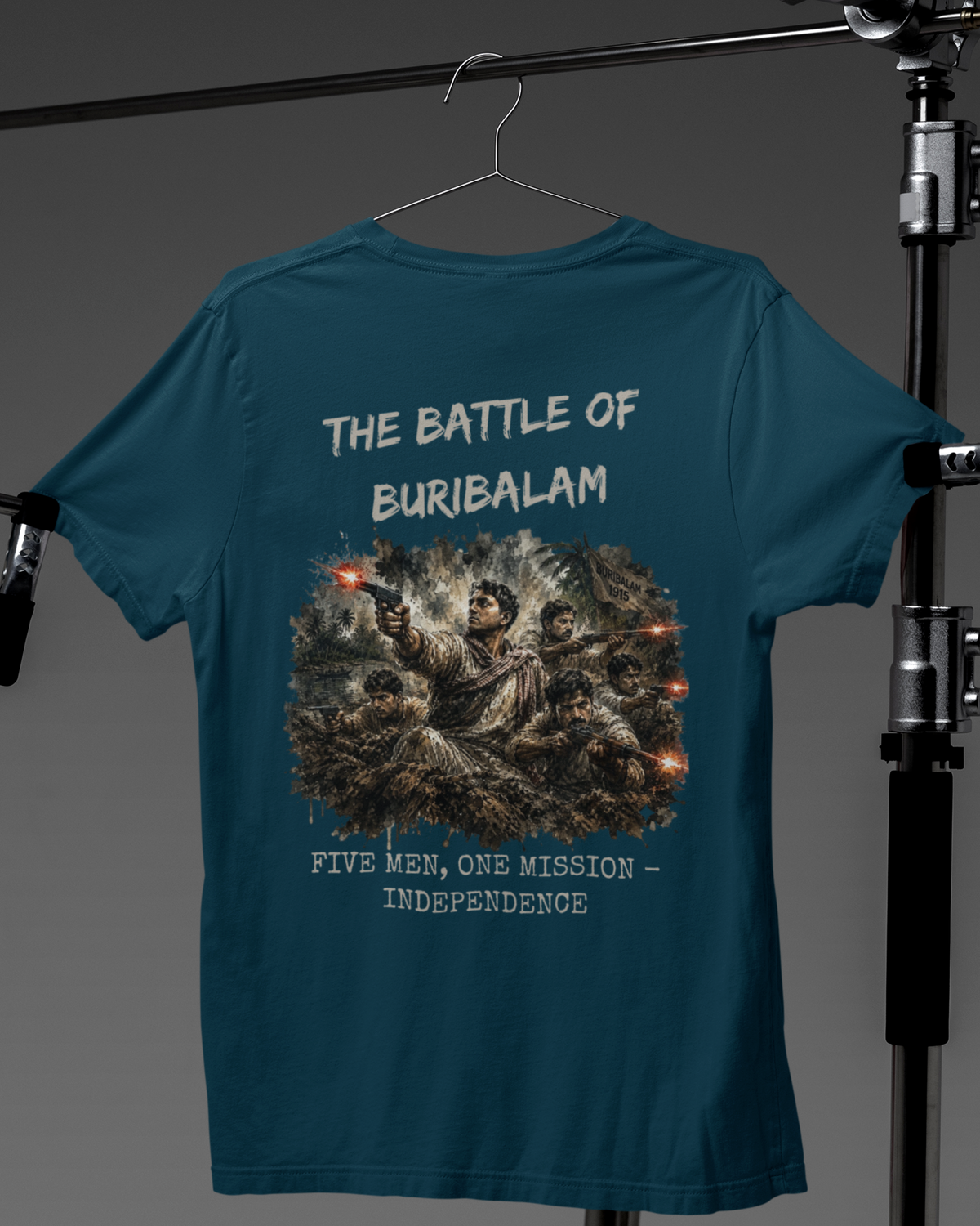 A teal blue graphic t-shirt hanging on a hanger against a dark gray studio backdrop, with a large back print reading 'THE BATTLE OF BURIBALAM' and 'FIVE MEN, ONE MISSION - INDEPENDENCE' around an illustration of soldiers.
