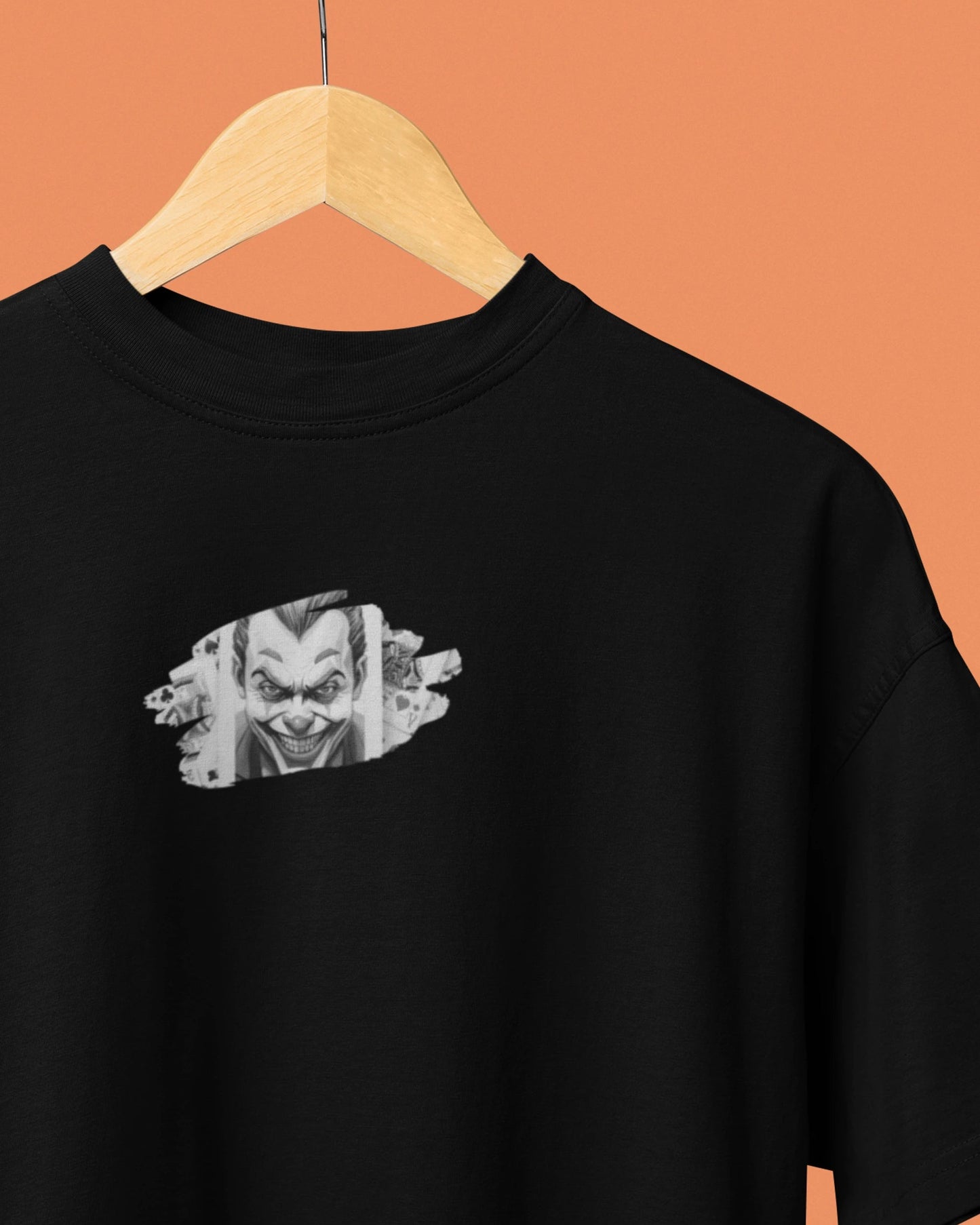 A black short-sleeve t-shirt hanging on a wooden hanger against a solid orange background. The shirt features a graphic print of a creepy clown face on the left chest, styled as if tearing through a piece of cardboard.