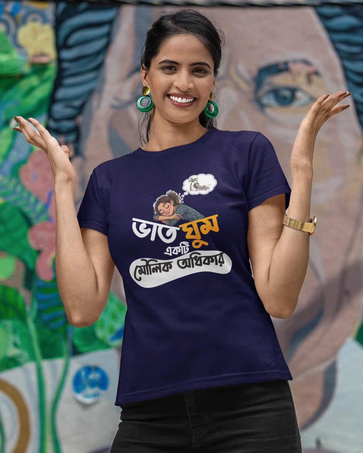 A woman posing in front of a colorful painted mural, wearing a navy blue graphic t-shirt with Bengali text and a cartoon illustration, paired with black pants.
