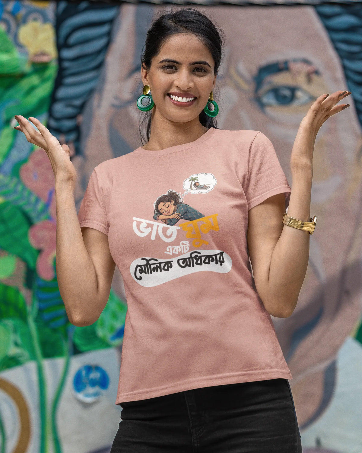 A woman posing in front of a colorful graffiti wall, wearing a dusty pink graphic t-shirt with Bengali text and a cartoon illustration, paired with black pants.