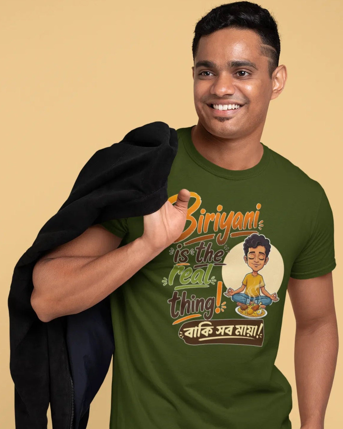A smiling man wearing an olive green graphic t-shirt with Bengali text and a cartoon illustration, holding a black jacket over his shoulder, set against a solid beige background.
