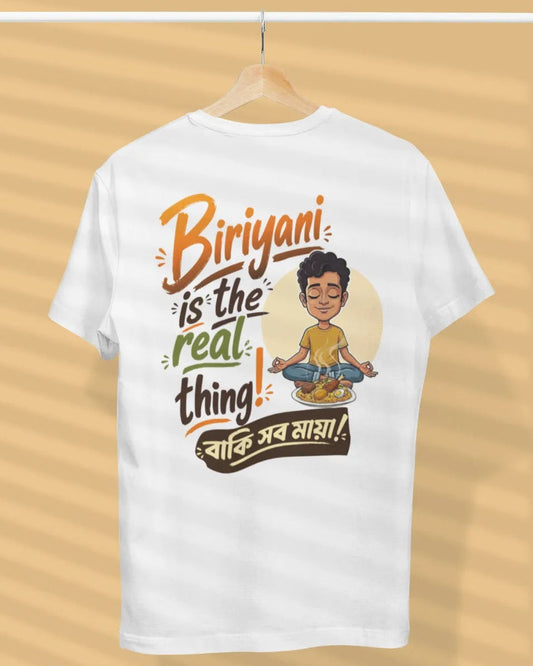 A white short-sleeve t-shirt hanging on a wooden hanger against a beige background with soft diagonal shadows. The back of the shirt features a colorful graphic with the text 'Biriyan! is the real thing!' and Hindi text, alongside a cartoon illustration of a man meditating.
