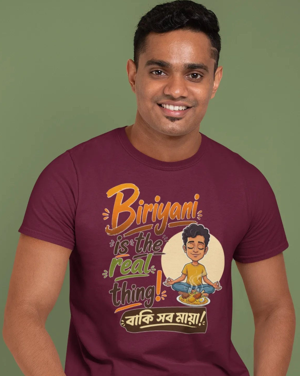 A man wearing a maroon graphic t-shirt and blue jeans, sitting on a wooden stool against a solid olive green background. The t-shirt features a colorful cartoon graphic and the text 'Biriyan! Is the real thing! वाकि सब लाया!'.