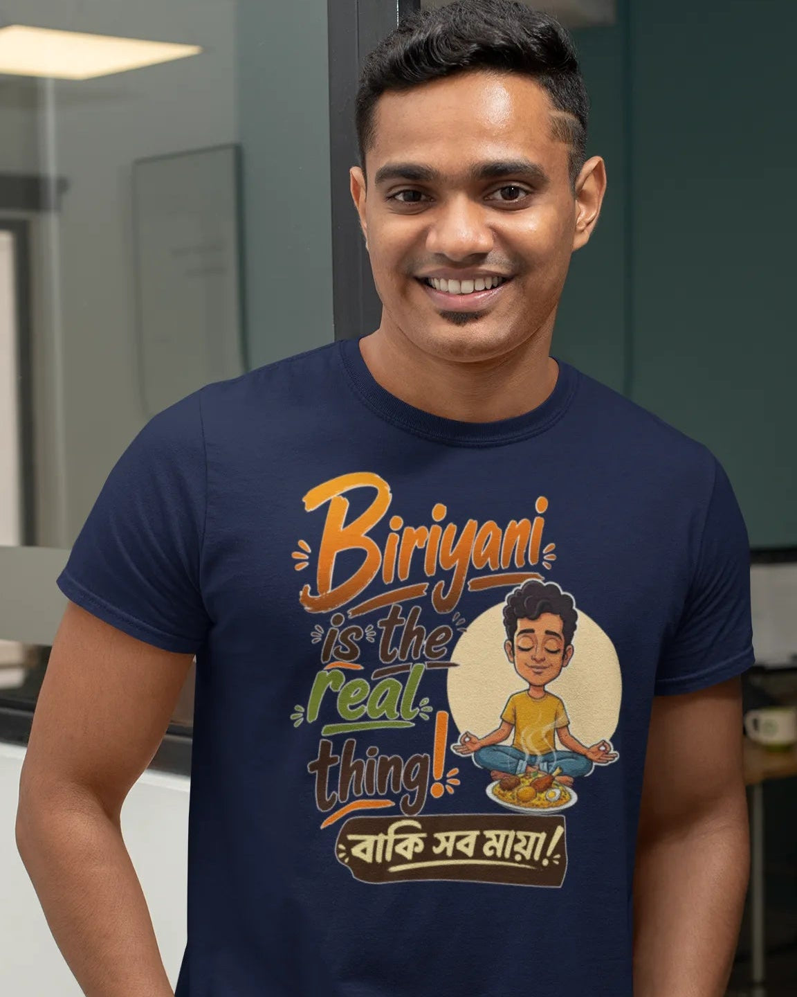 A man wearing a navy blue graphic t-shirt with a cartoon illustration and the text 'Biriyan is the real thing! বাংলা সব সাফাই!' on the front, standing in an office environment.