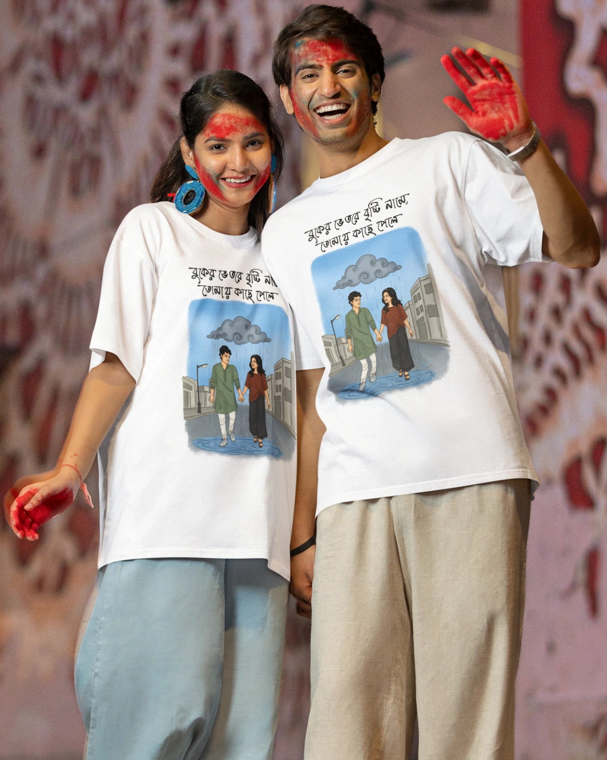 A man and a woman standing together and holding hands, both wearing matching white graphic t-shirts with a colorful illustration and Bengali text. The woman is wearing light blue wide-leg pants and lime green sneakers, while the man is wearing beige wide-leg pants and green sneakers.
