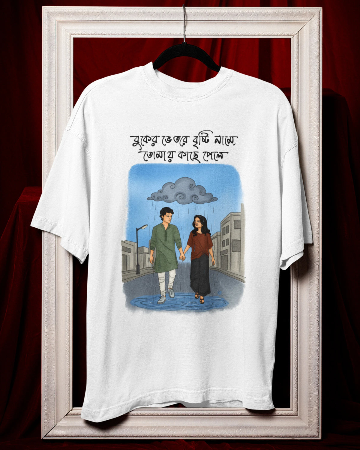 A white short-sleeve graphic T-shirt hanging on a black hanger inside a large white picture frame, set against a dark red draped fabric background. The shirt features a central illustration of a man and woman walking in the rain under a gray cloud, with Bengali text above the graphic.