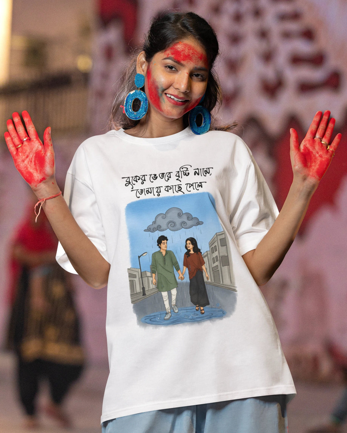 A woman wearing a white graphic t-shirt with Bengali text and an illustration, posing with red paint on her hands and face against a blurred street backdrop.