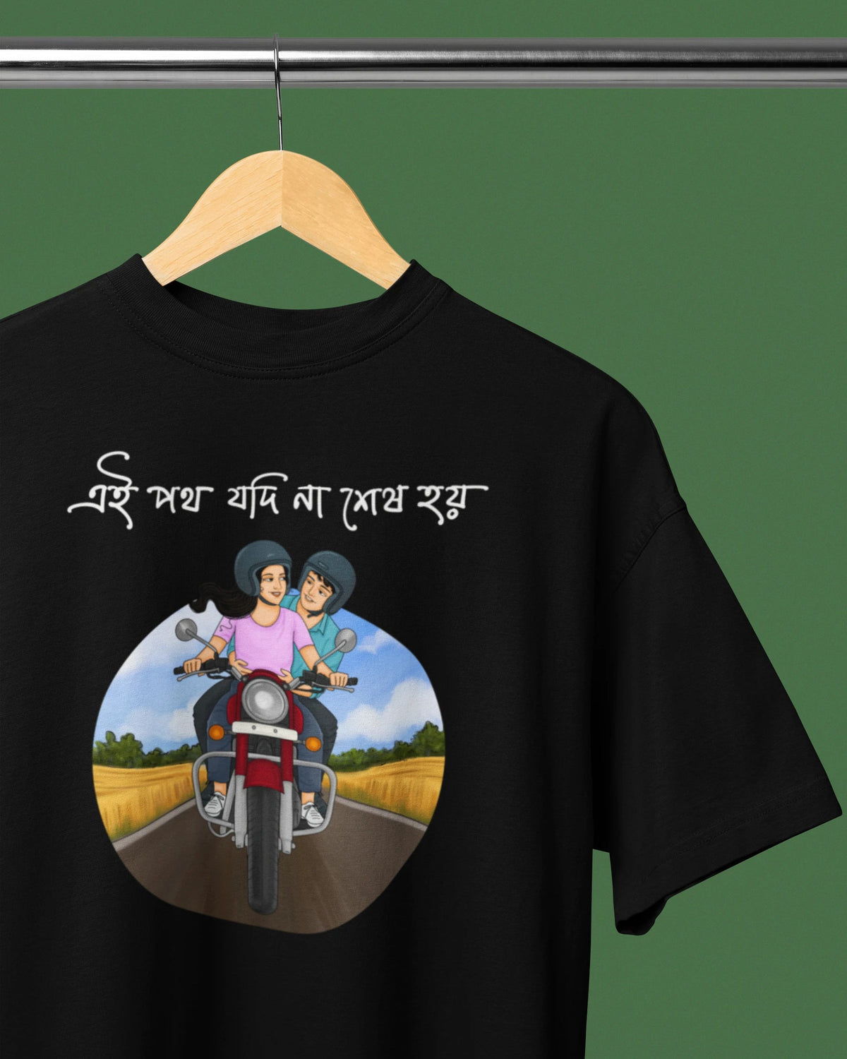 A black short-sleeve t-shirt hanging on a wooden hanger against a solid green background. The shirt features white Bengali text above a circular graphic illustration of a couple riding a motorcycle on a country road.