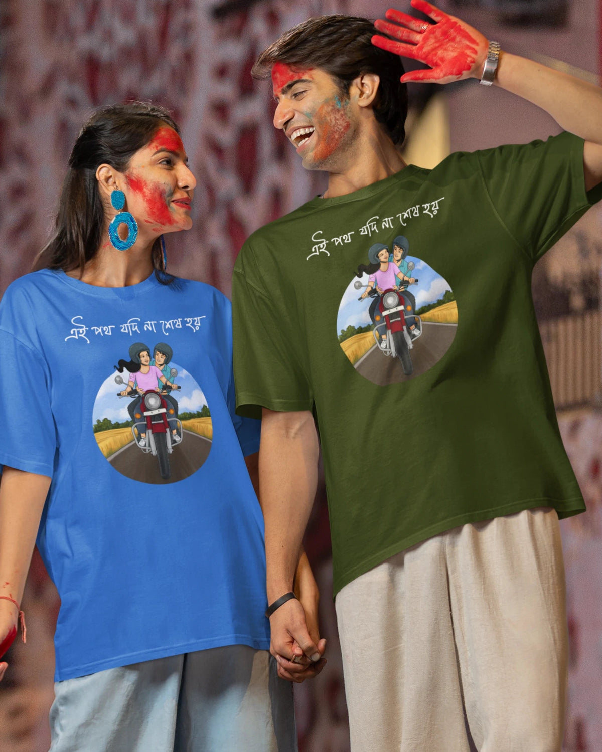A man and a woman holding hands, wearing matching graphic t-shirts with a Holi-themed circular print and Gujarati text. The woman wears a blue t-shirt and light blue jeans, while the man wears an olive green t-shirt and beige pants.