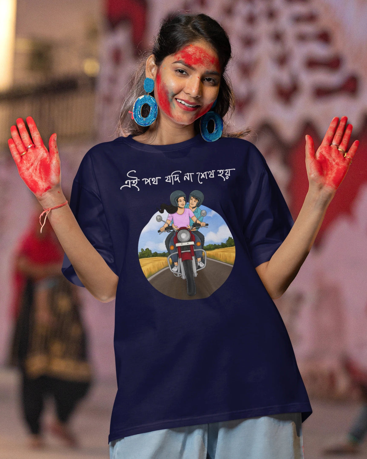 A woman wearing a navy blue graphic t-shirt with a circular print of a girl on a motorcycle and Devanagari text, posing with red face and hand paint in an outdoor street setting.