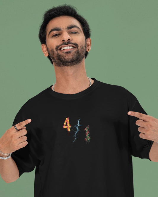 A smiling man wearing a black t-shirt with a colorful graphic on the chest, pointing at the design with both hands against a solid green background.