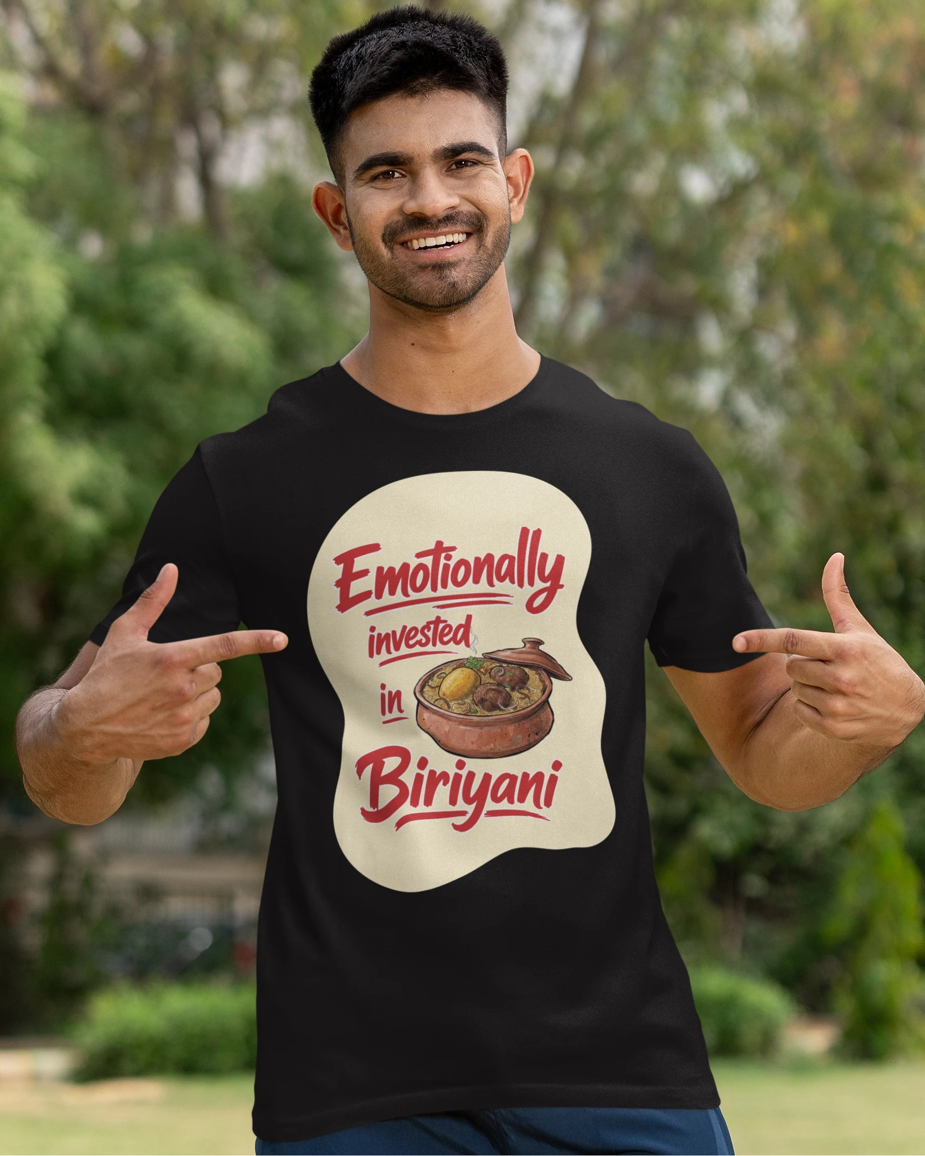 A man wearing a black graphic t-shirt with a 'Emotionally invested in Biriyani' print, posing outdoors with a cricket bat against a blurred green background.