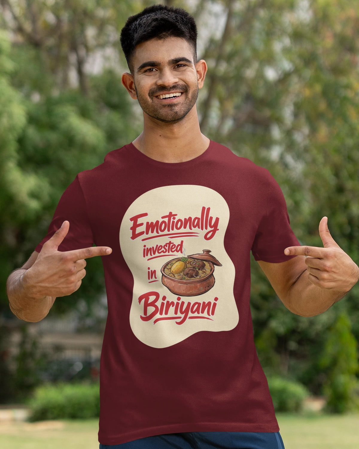 A man wearing a maroon t-shirt with a graphic print that reads 'Emotionally invested in Biriyani' in red text on a cream-colored splash shape. He is posing outdoors with a cricket bat in a grassy park setting.